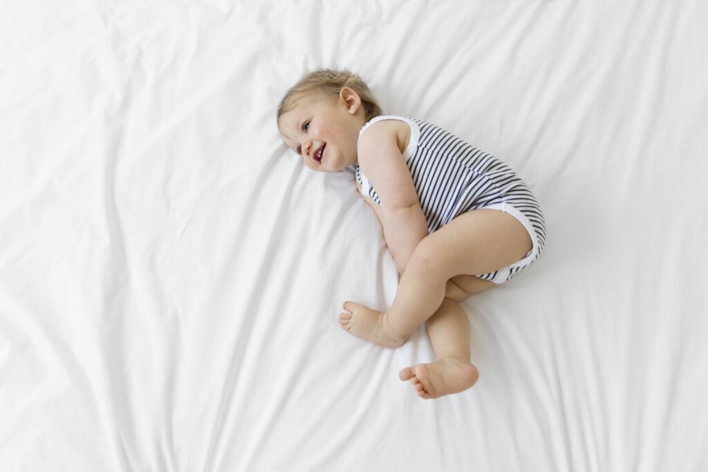 High Angle View Of Baby Boy Wearing Striped Onesie Lying On Bed With White Duvet.